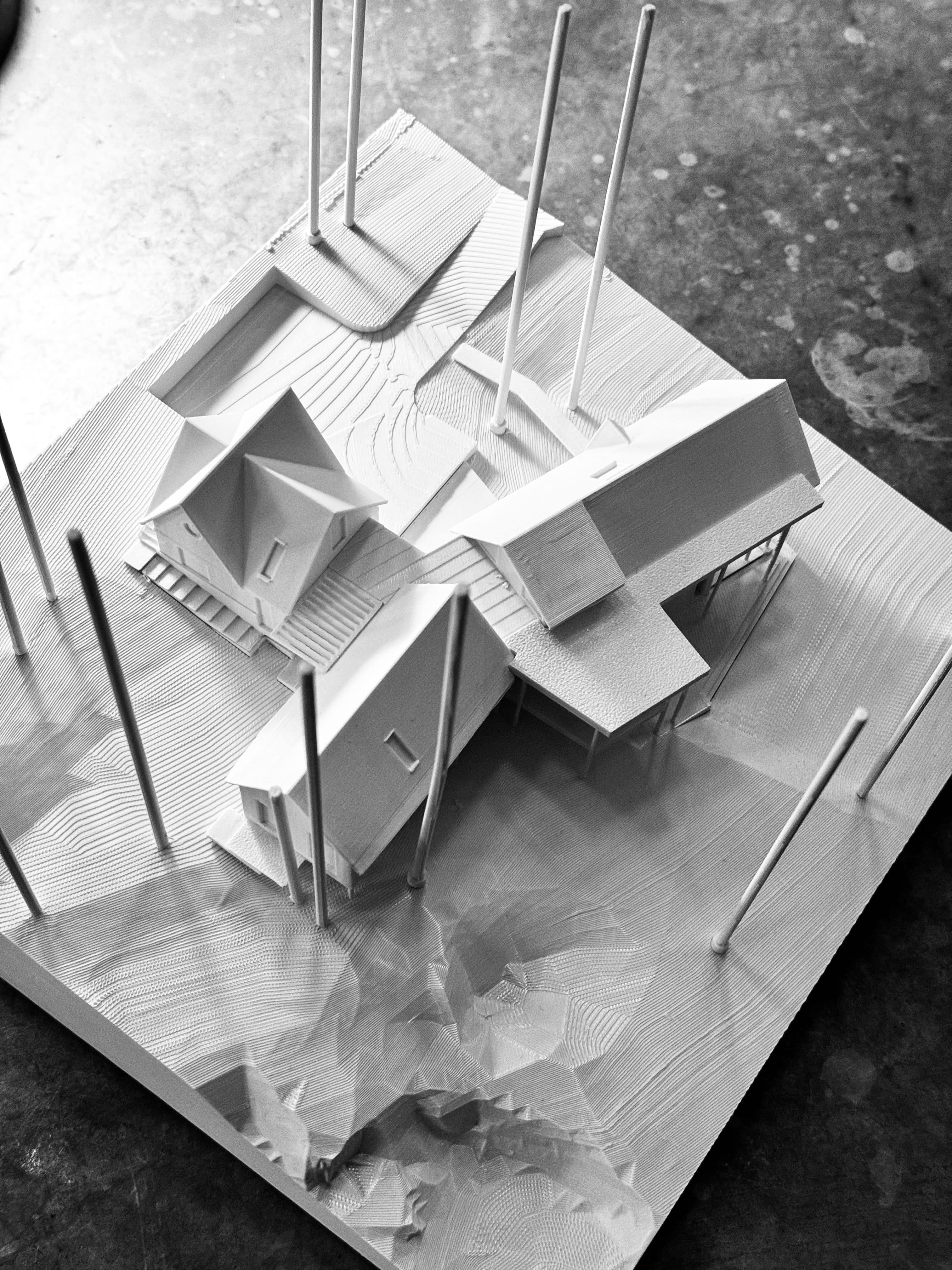 A white study model of Canal House's stacked volumes on point supports, set into a carved topographic base representing the sloped, wooded site.