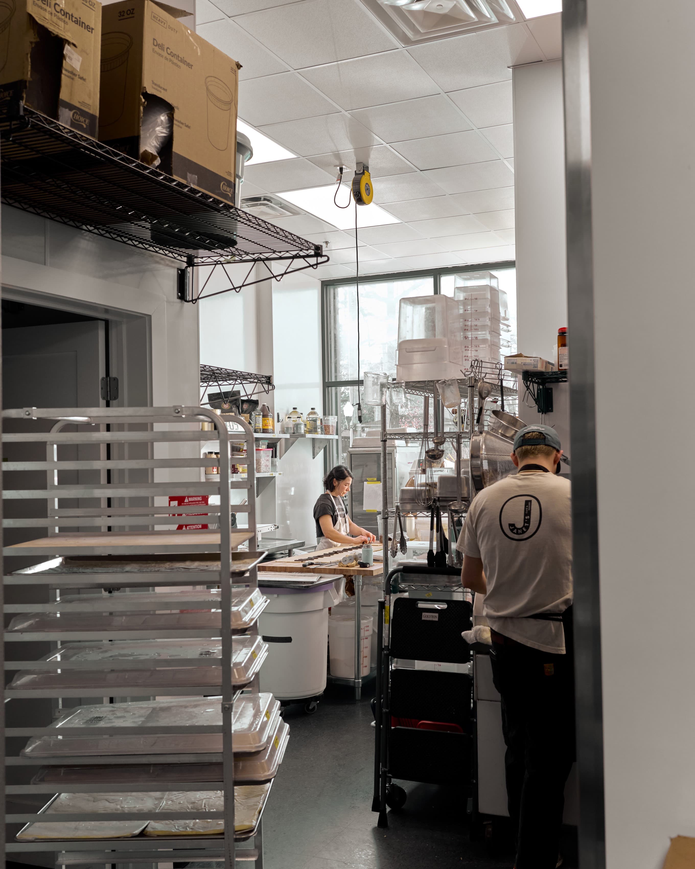 The bakery's production kitchen seen through the doorway from the front of the house, a baker at a wood bench beneath a tall window, wire shelving and a rolling tray rack around.