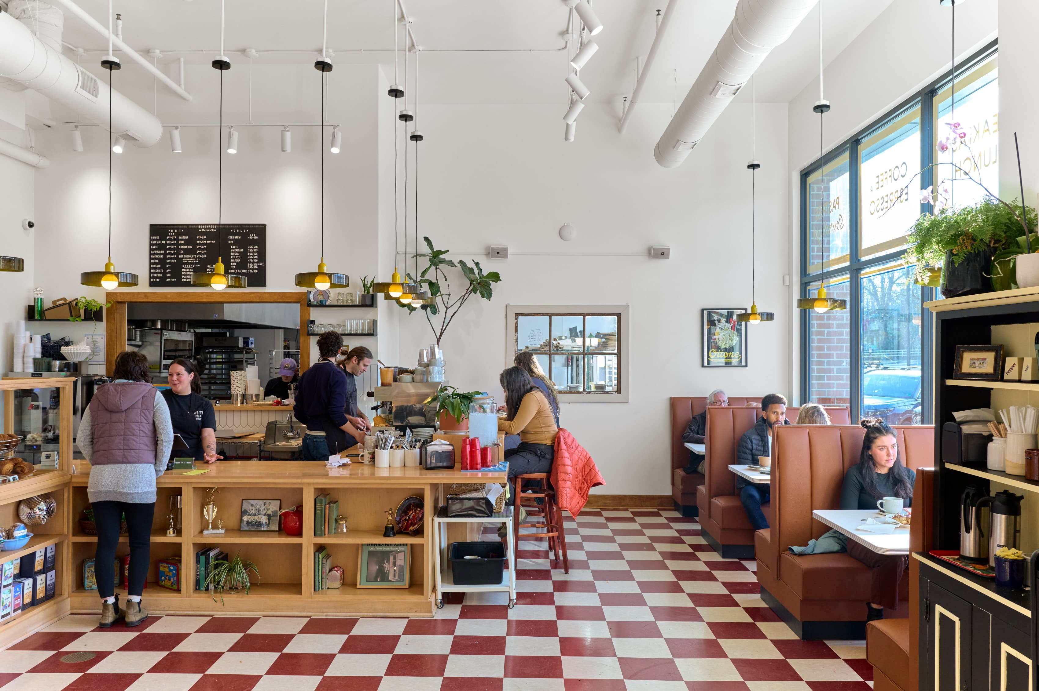 The full counter run and kitchen pass beneath exposed, painted-white ductwork and pendant lighting, seating booths and storefront glazing at right, checkerboard floor underfoot.