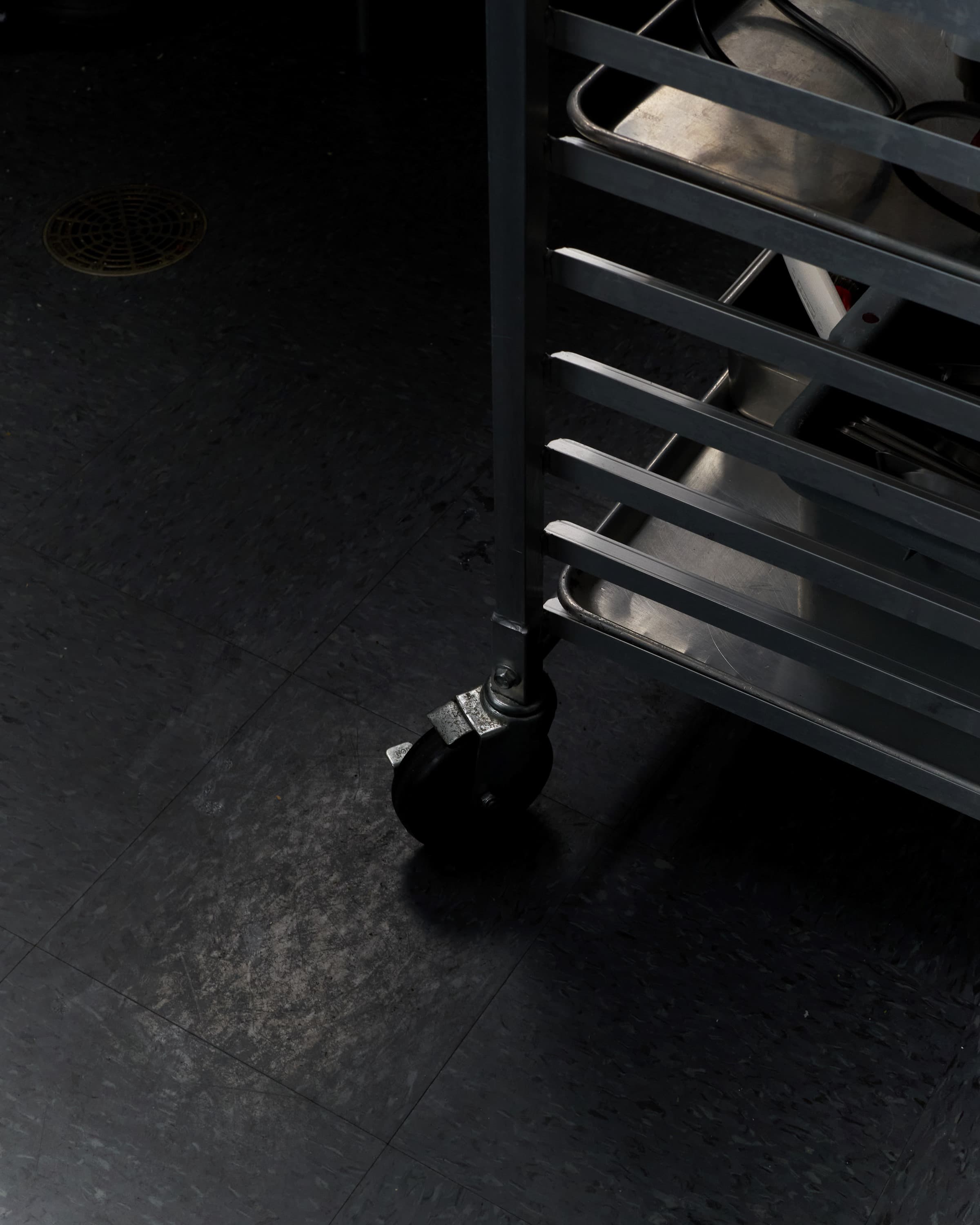 A stainless steel rolling rack's caster wheel on the bakery's dark tile floor, a floor drain beyond.