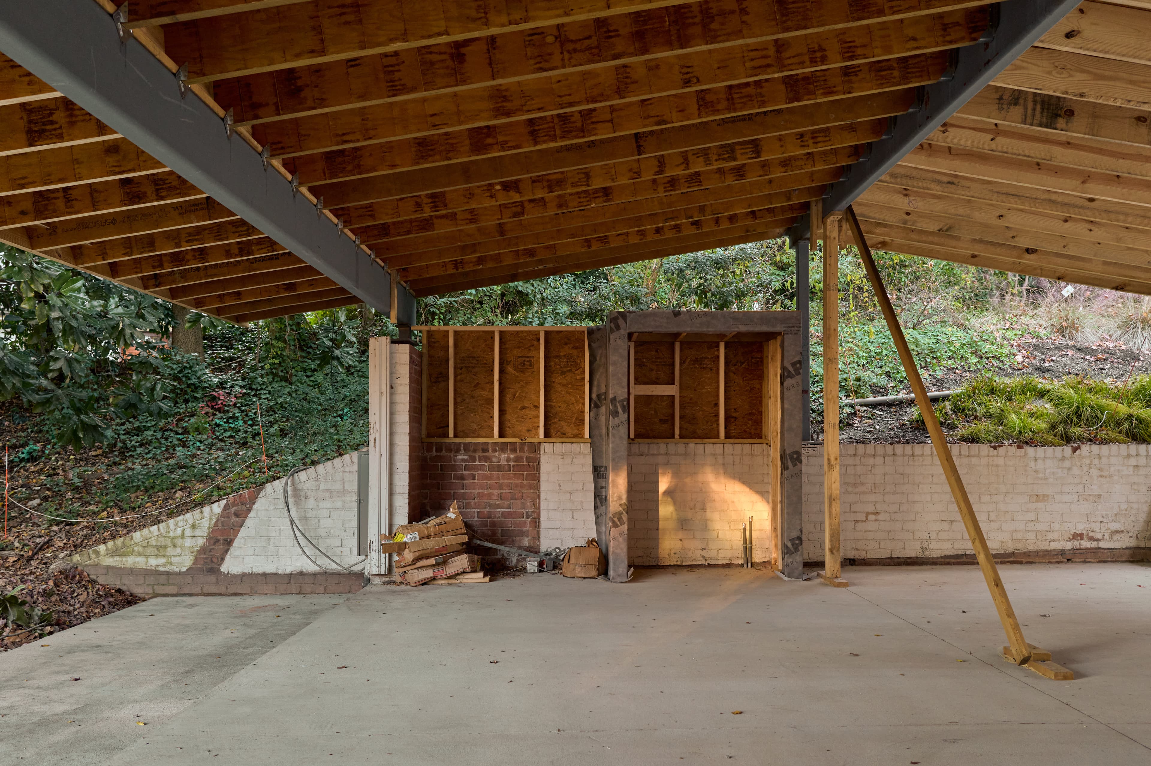 Wide view beneath the new carport's steel-and-wood roof structure, framed openings wrapped in preparation for cladding, existing brick wall beyond.
