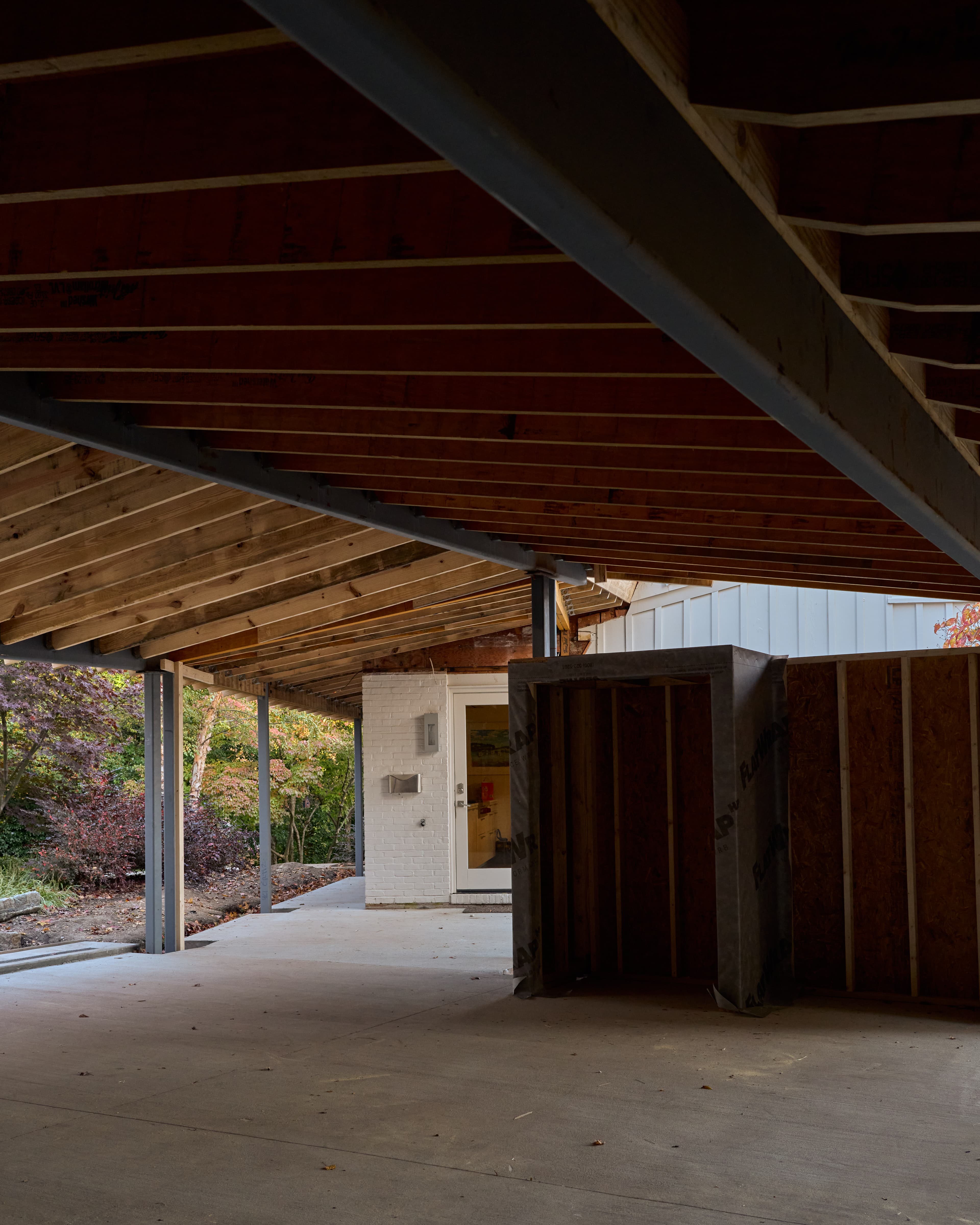 A new steel-and-wood carport roof structure under construction, meeting the existing painted-brick house at a door and brick pier.