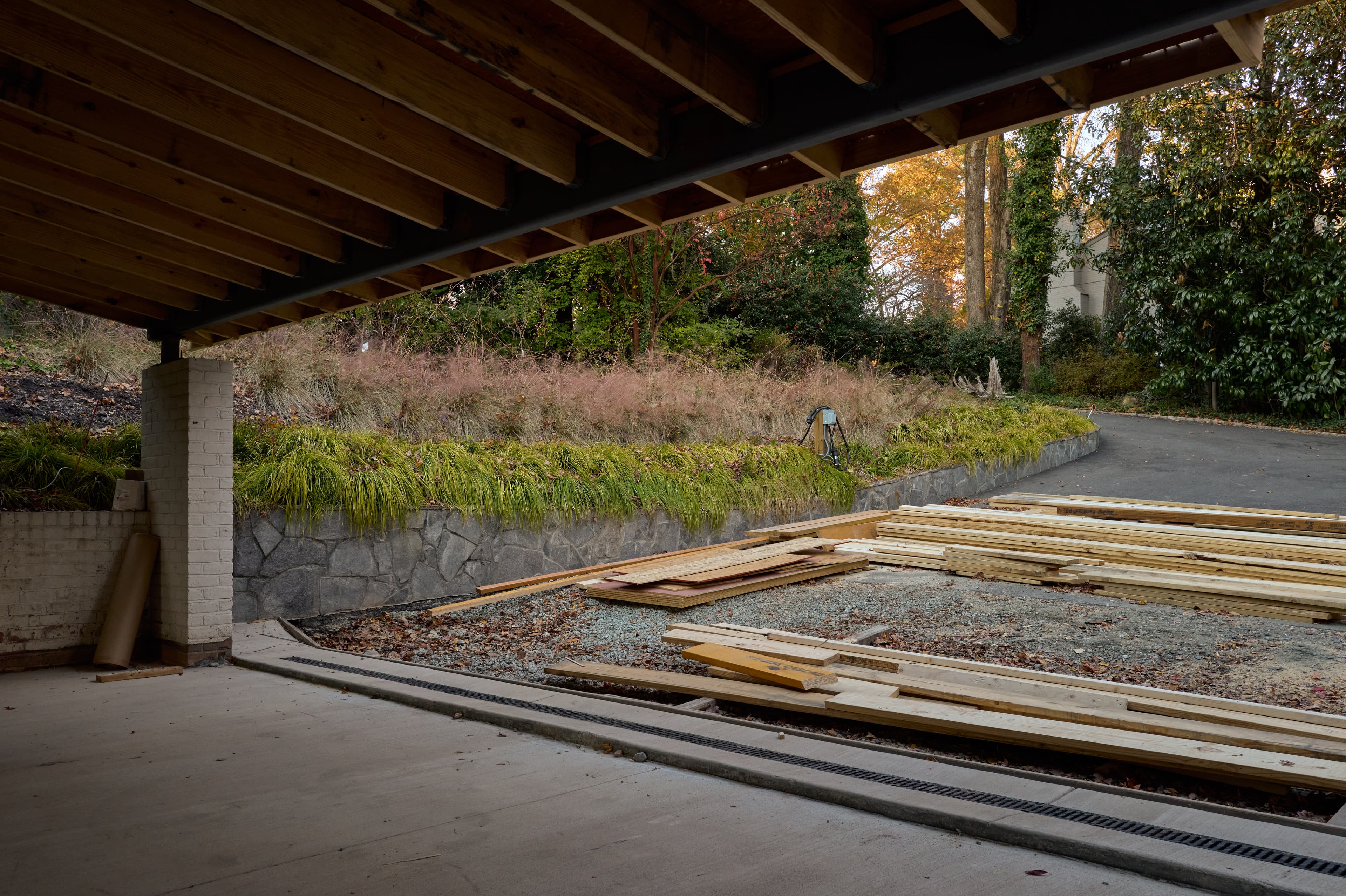 The new carport's steel-and-wood roof structure seen from beneath, open toward the driveway, retaining wall, and planted slope beyond.