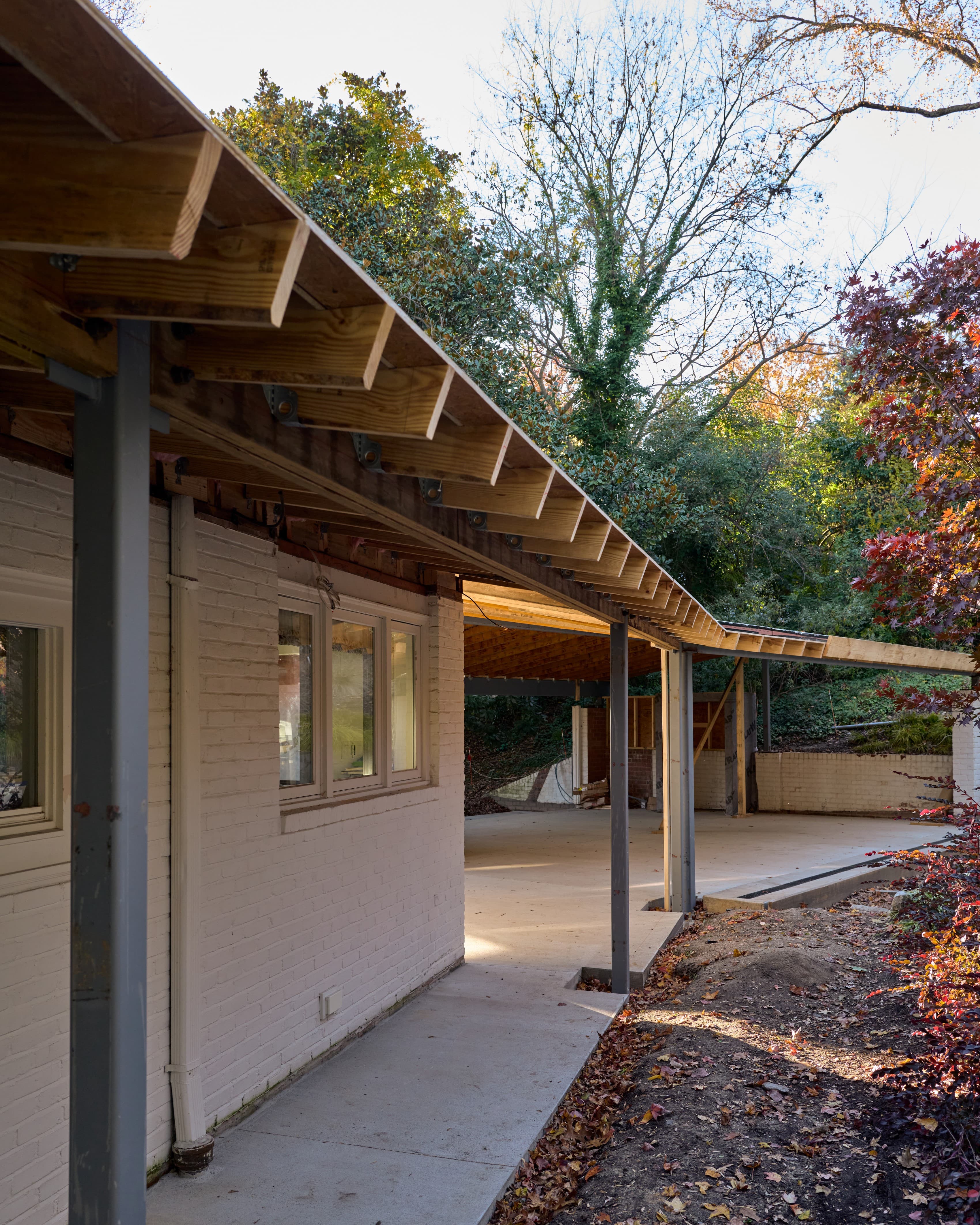 The existing painted-brick house at Tempsford Residence with a new Accoya-lined roof plane on steel columns extending from it toward a new carport structure, under construction.