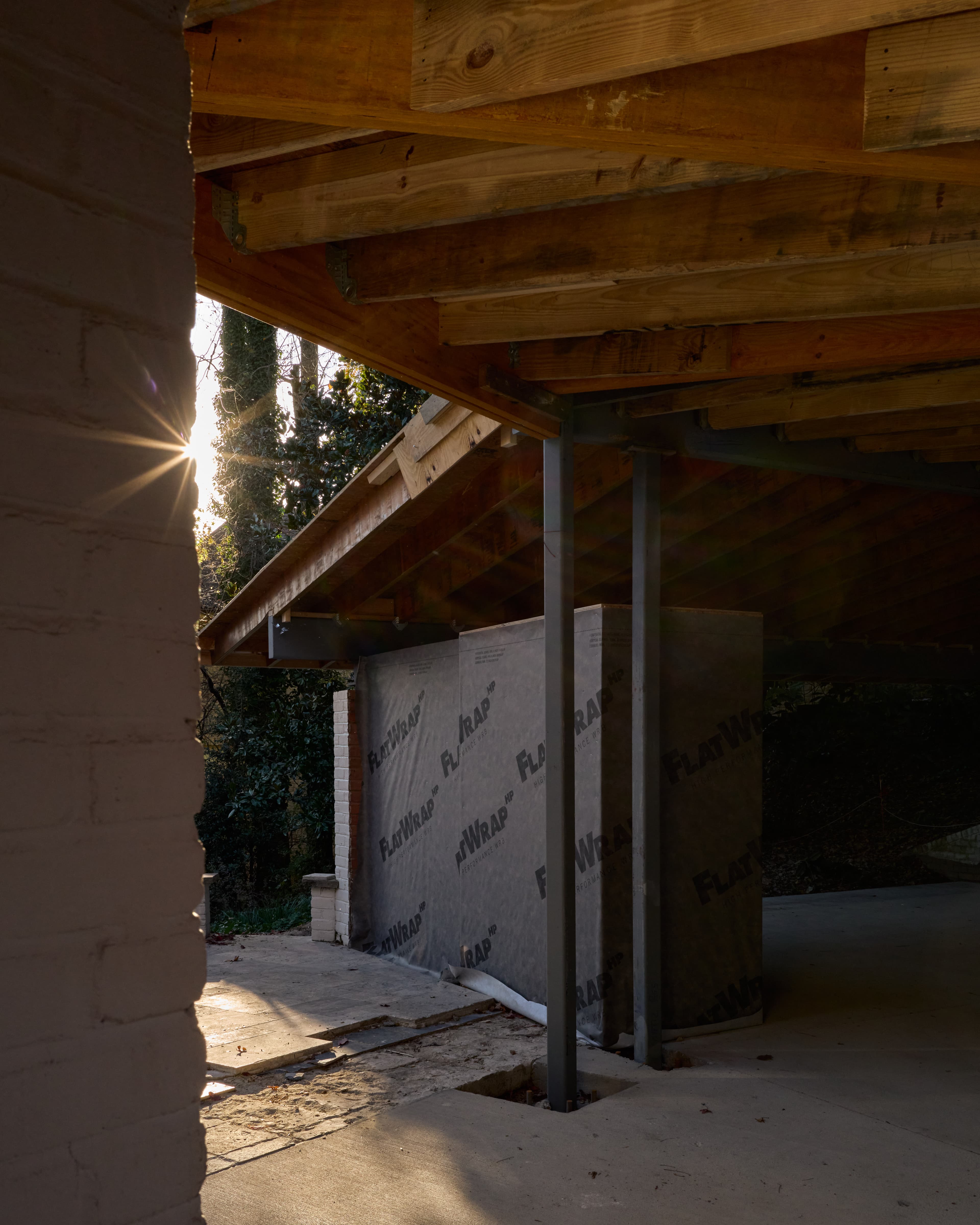 Low sun through the new structure's timber framing, a weather-resistive-barrier-wrapped wall and steel column in the foreground, existing brick pier beyond.